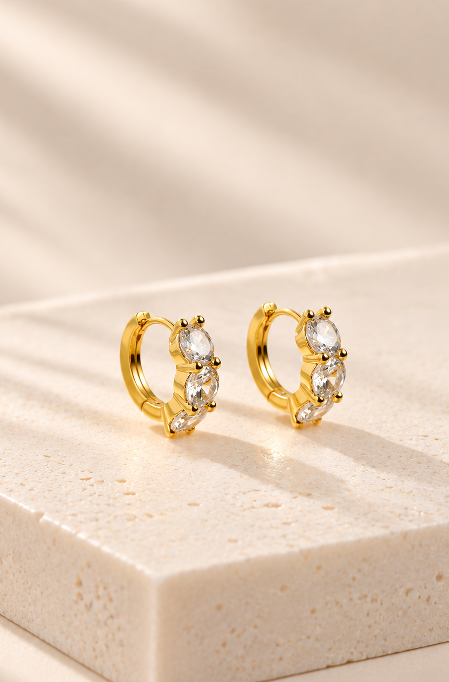 A pair of gold huggie hoop earrings with clear marquise-cut gemstones, displayed on a textured beige stone block with soft, warm shadows.