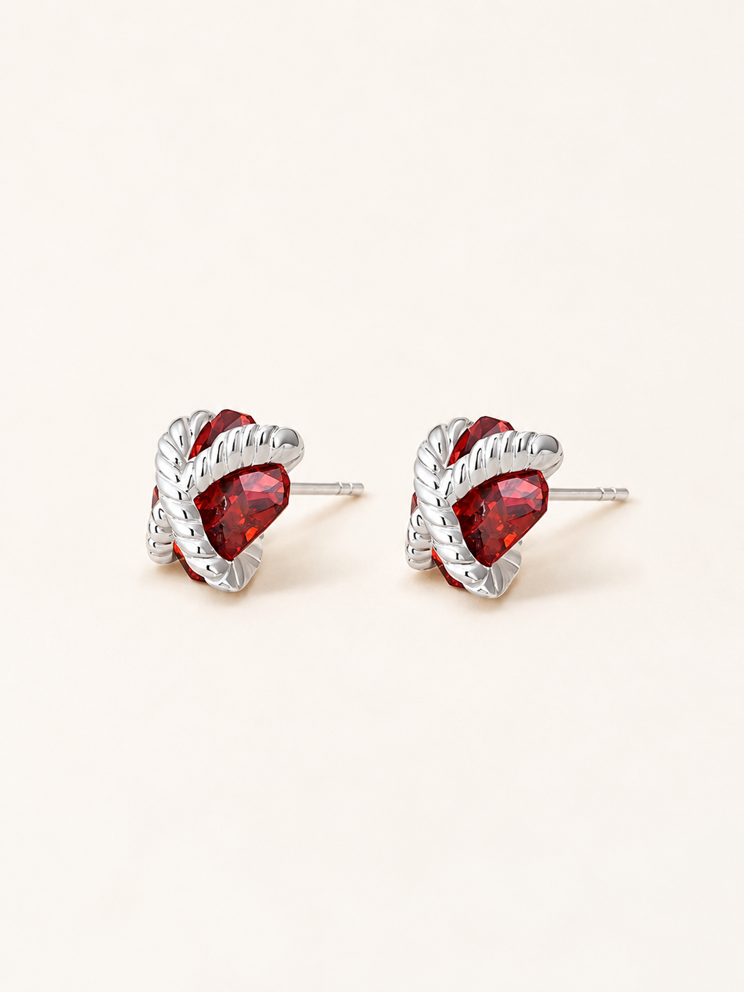 A pair of silver-toned twisted rope stud earrings with faceted red heart-shaped crystals, displayed on a clean, light cream background.