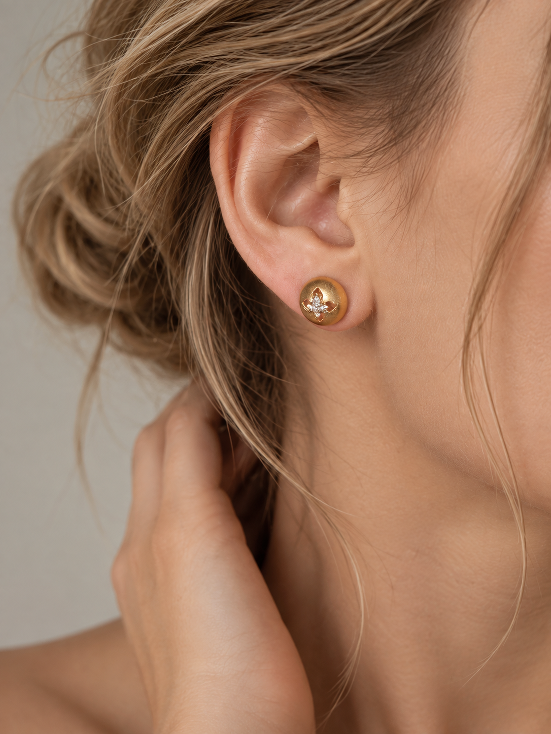 A close-up of a woman's ear wearing a small gold stud earring with a clear floral crystal, set against a soft neutral background.
