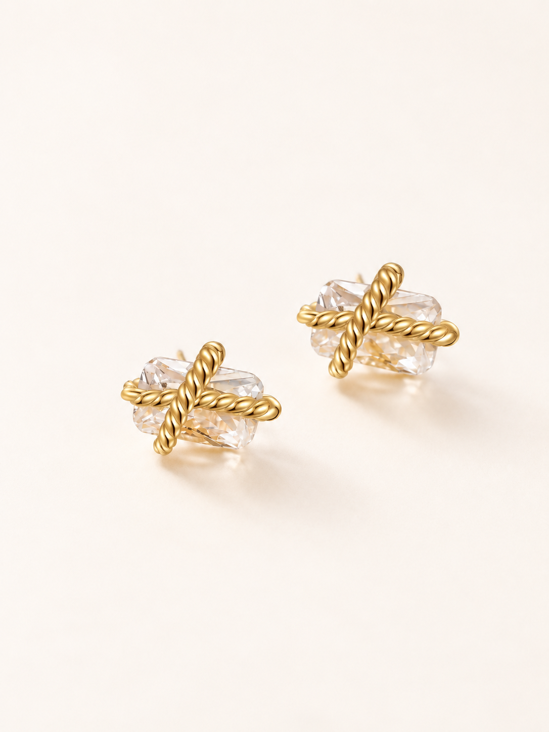 A pair of gold stud earrings featuring a twisted rope design wrapping around a clear faceted crystal, set against a warm off-white background.