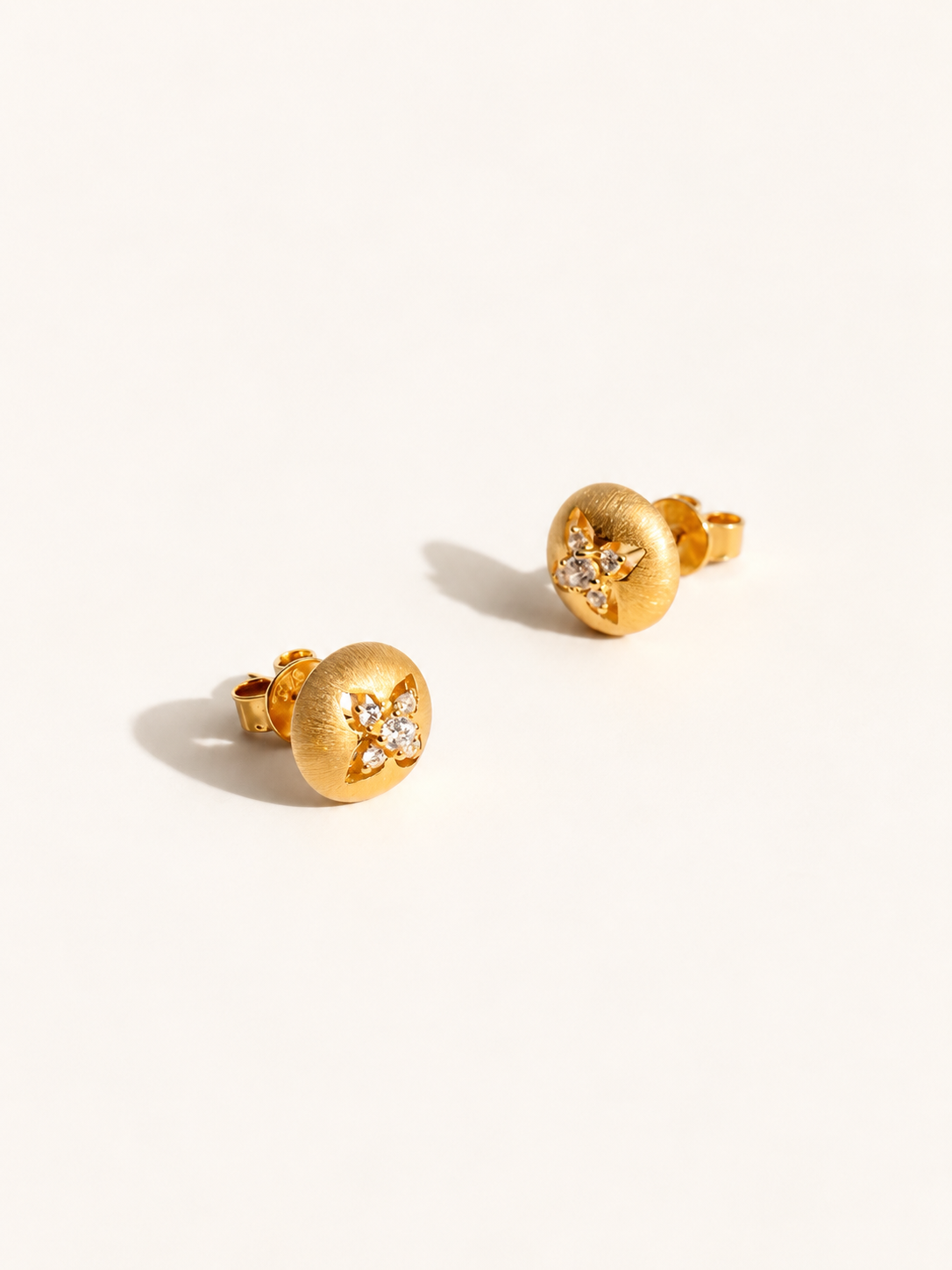 A pair of gold stud earrings with a textured circular face and a small cluster of clear stones in a floral or starburst motif, photographed on a clean off-white background.