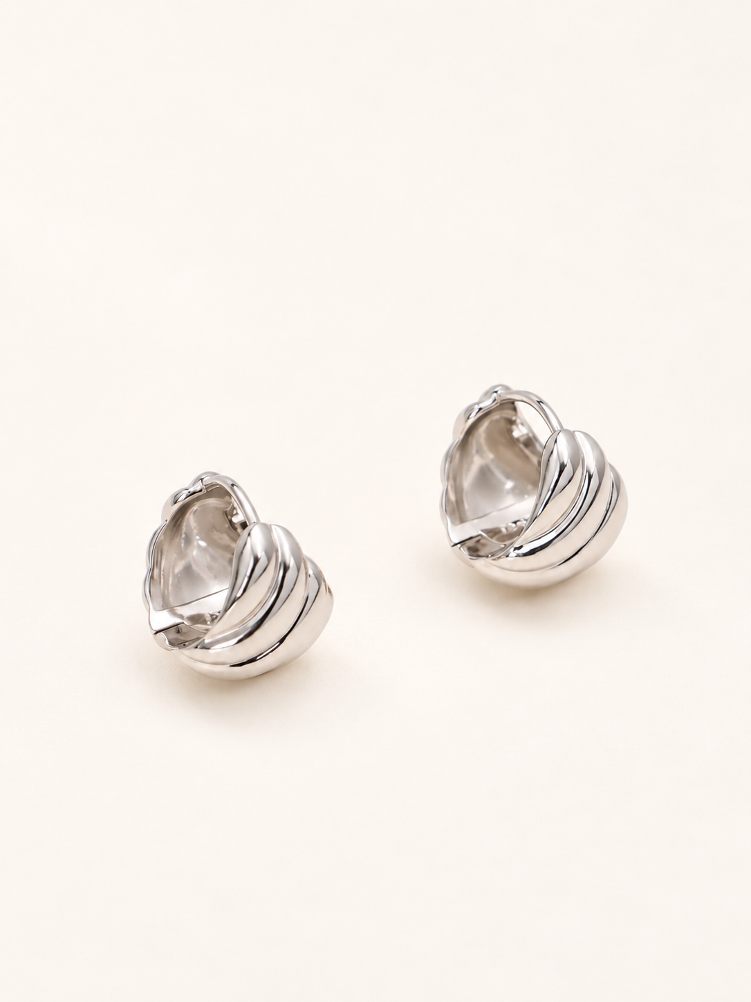 A pair of silver-toned, sculptural stud earrings with a ribbed, organic shell-like design, displayed on a clean, light cream background.