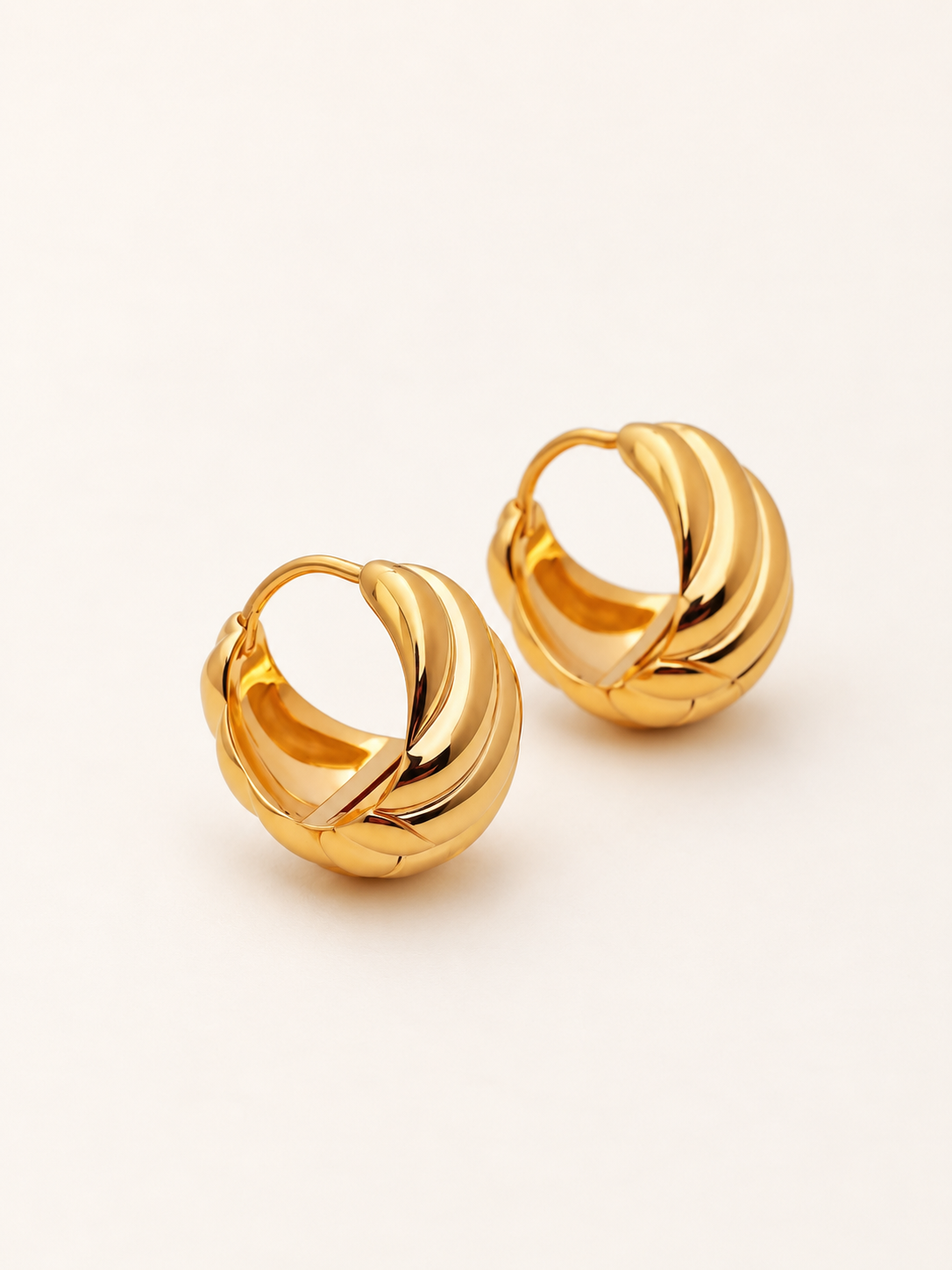A pair of gold huggie hoop earrings with a twisted, layered design, photographed on a clean, light off-white background.