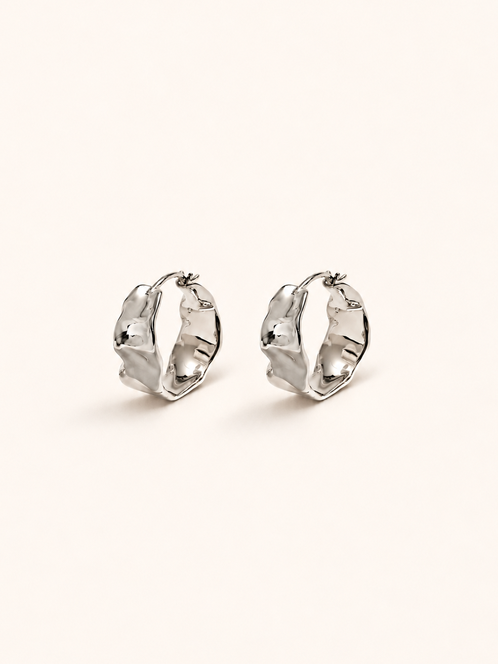 A pair of silver-toned textured hoop earrings on a plain, warm off-white background.