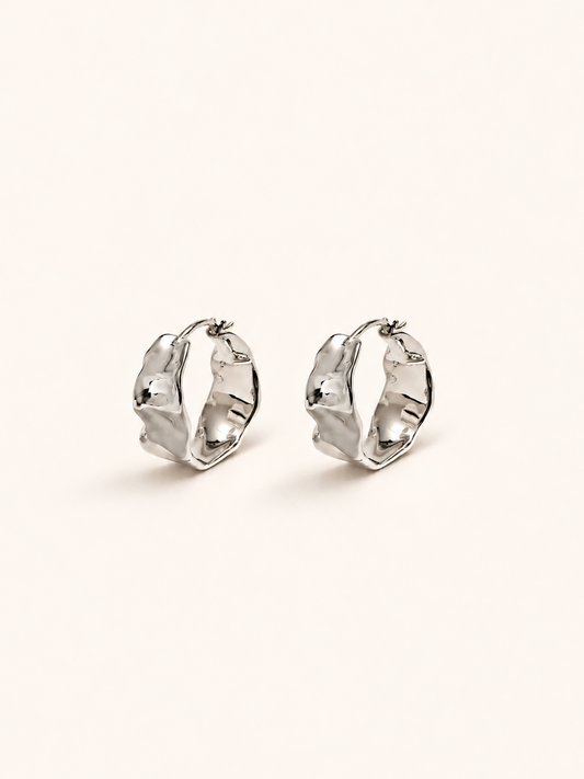 A pair of silver-toned textured hoop earrings on a plain, warm off-white background.