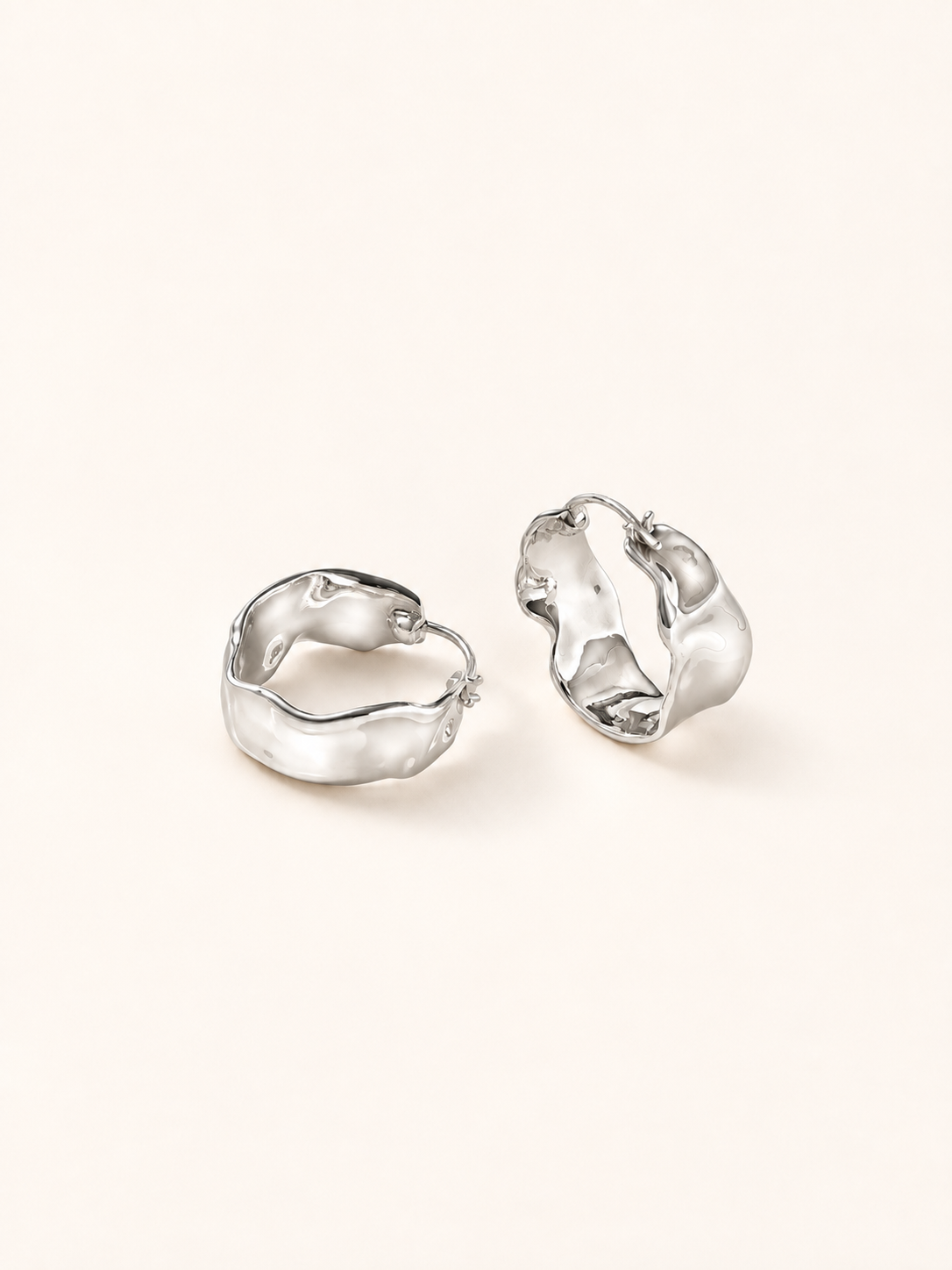 A pair of silver-toned, organic-shaped hoop earrings with a textured, wavy surface, displayed on a clean, solid off-white background.